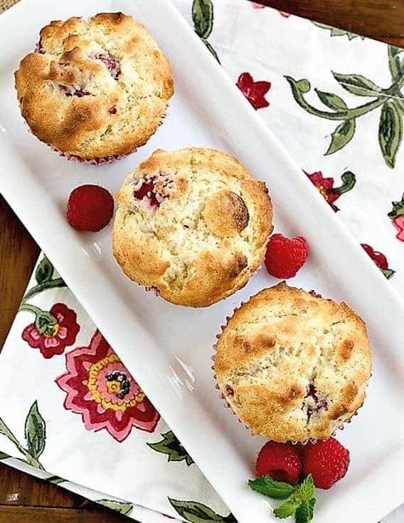 Overhead view of 3 Raspberry White Chocolate Muffins on a rectangular tray with fresh raspberries and a sprig of mint.