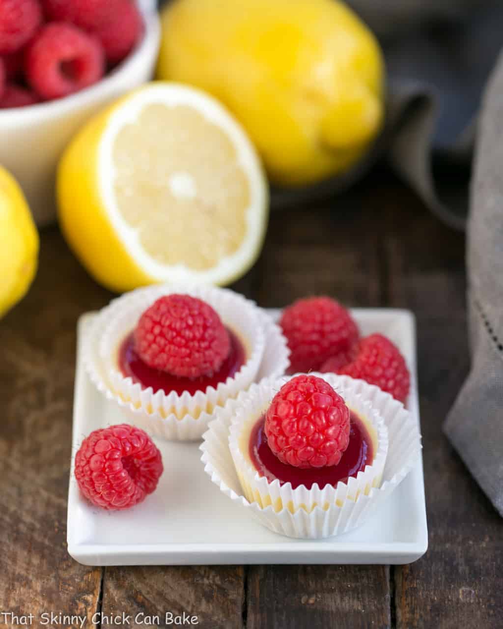 Two raspberry topped cheesecake bites on a small white plate with raspberry garnishes.