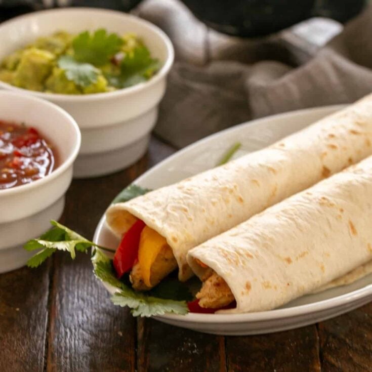 Two chicken fajitas on a white plate in front of bowls of guacamole and salsa.